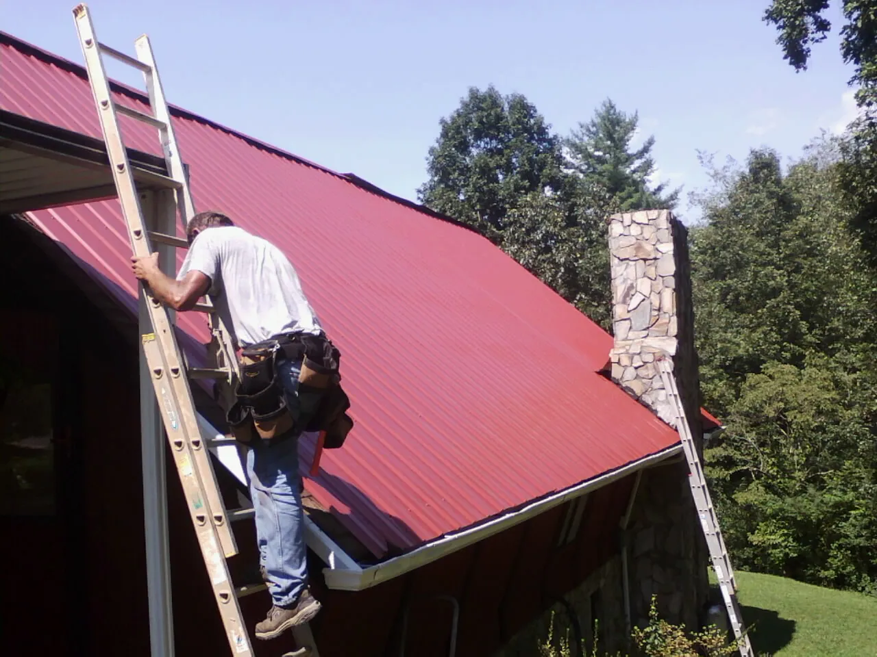 Roofer on a ladder beside a red metal roof and stone chimney — Soffit & Fascia Repair by Roofing Brothers in Island